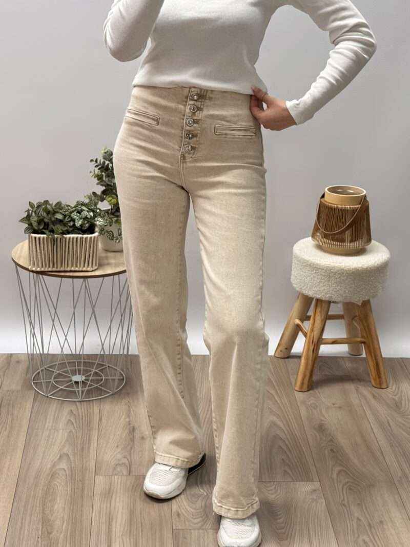 jean bootcut beige taille haute avec fermeture à boutons apparents, porté avec un haut ajusté blanc et des baskets beiges. Couple fluide et évasée, idéale pour allonger la silhouette. Photo prise dans un cadre minimaliste avec une touche naturelle.