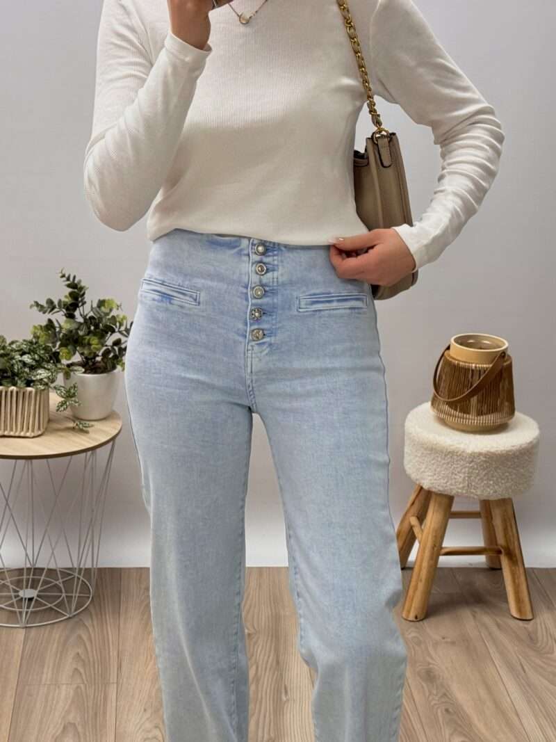 jean bootcut bleu clair taille haute avec fermeture à boutons apparents, porté avec un haut ajusté blanc et des baskets beiges. Couple fluide et évasée, idéale pour allonger la silhouette. Photo prise dans un cadre minimaliste avec une touche naturelle.