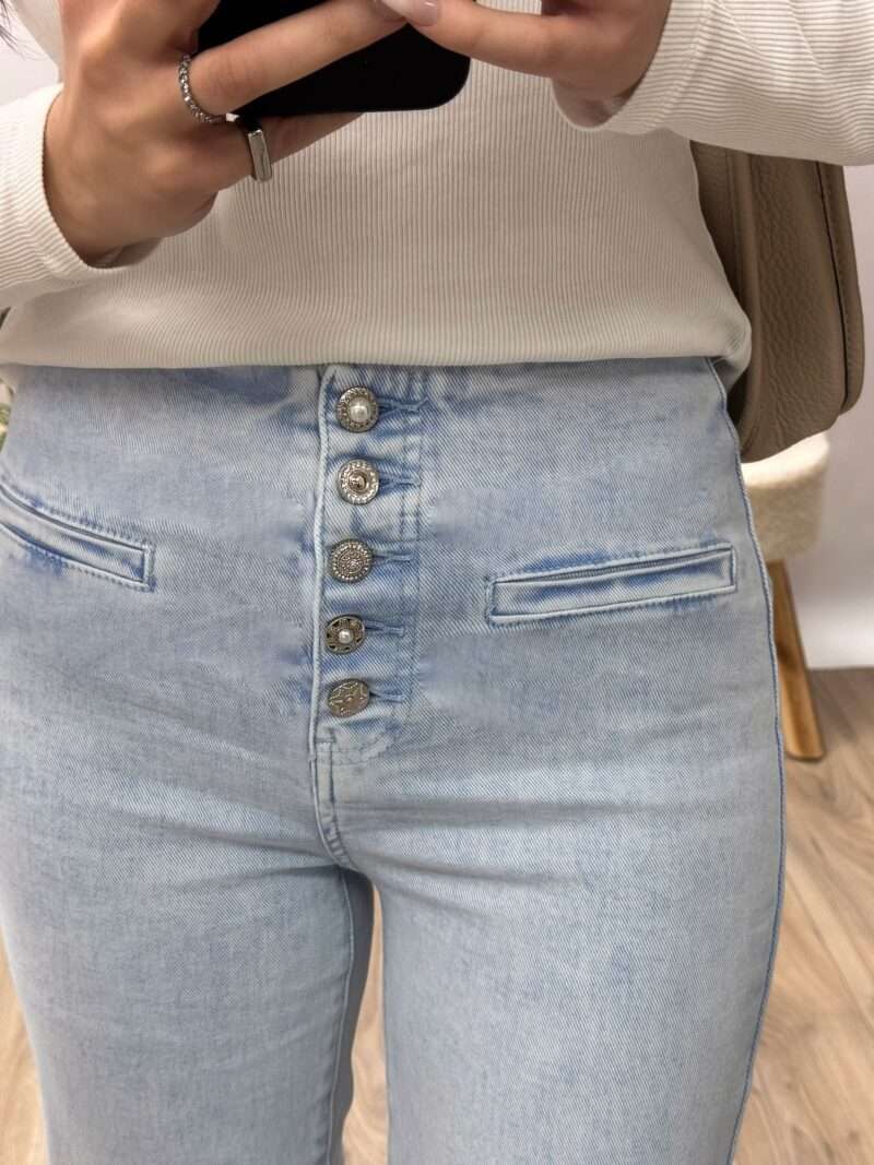 jean bootcut bleu clair taille haute avec fermeture à boutons apparents, porté avec un haut ajusté blanc et des baskets beiges. Couple fluide et évasée, idéale pour allonger la silhouette. Photo prise dans un cadre minimaliste avec une touche naturelle.