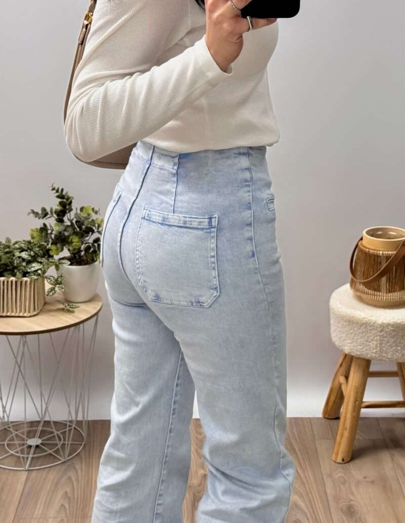 jean bootcut bleu clair taille haute avec fermeture à boutons apparents, porté avec un haut ajusté blanc et des baskets beiges. Couple fluide et évasée, idéale pour allonger la silhouette. Photo prise dans un cadre minimaliste avec une touche naturelle.