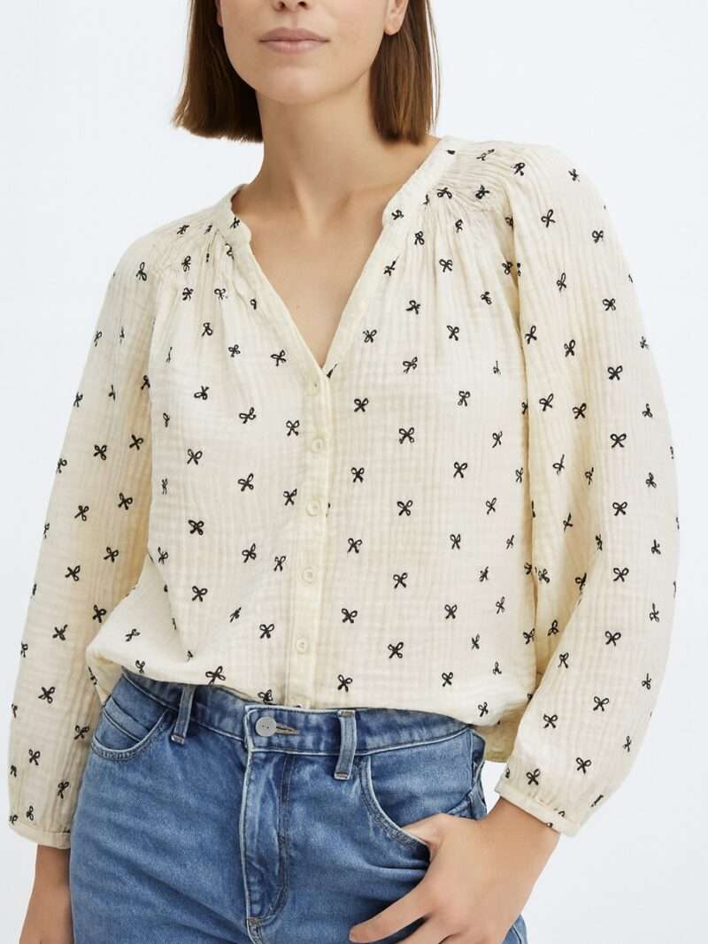 Femme portant blouse crème col V à petits nœuds noirs brodés, tissu texturé.