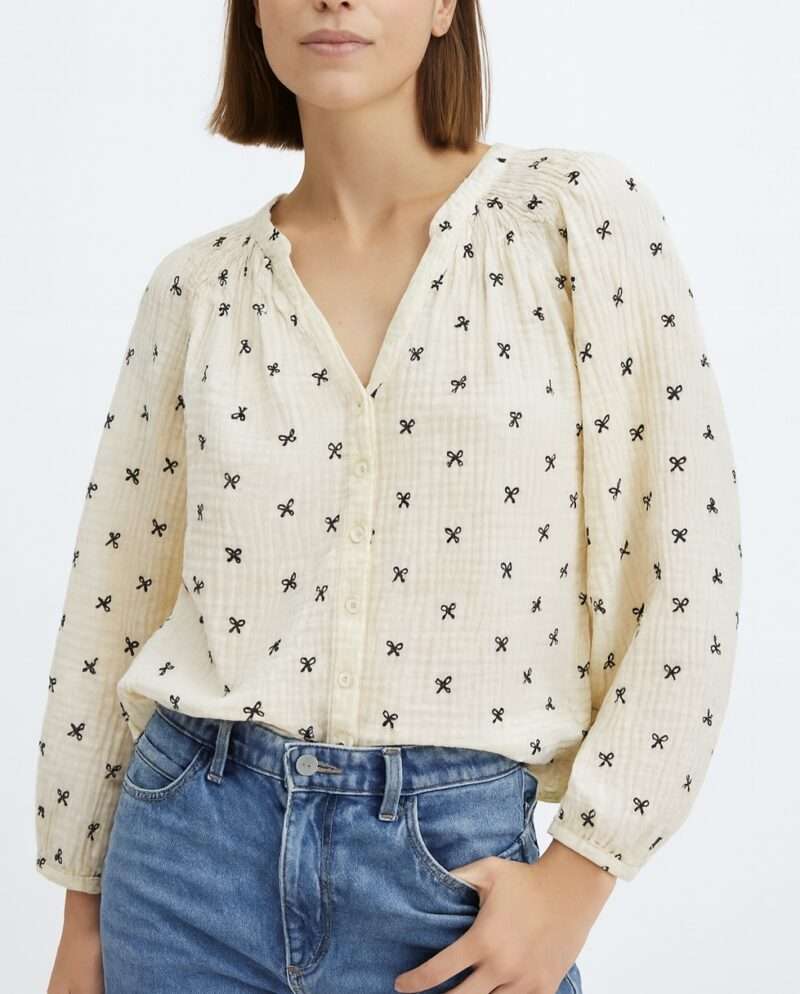 Femme portant blouse crème col V à petits nœuds noirs brodés, tissu texturé.